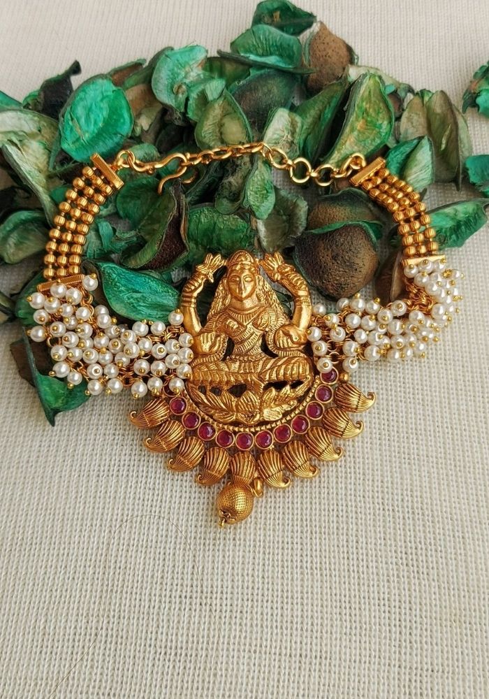 Elegant gold and pearl Laxmi Waaki/Bajuband against a backdrop of green leaves, showcasing a detailed Goddess Laxmi pendant in the center of the ornate jewelry piece from Mugdha Jewellery Store.