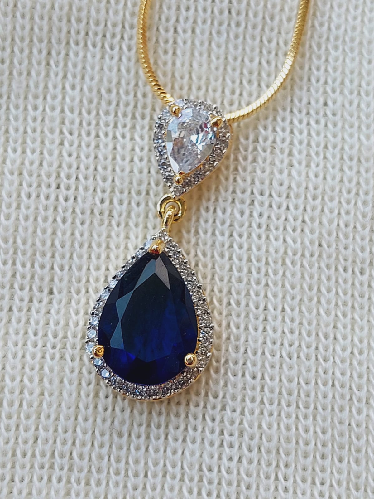 Elegant contemporary dark blue glass stone pendant on a gold chain. The pendant features a teardrop-shaped dark blue gemstone surrounded by a halo of sparkling clear stones, creating a sophisticated and luxurious look.