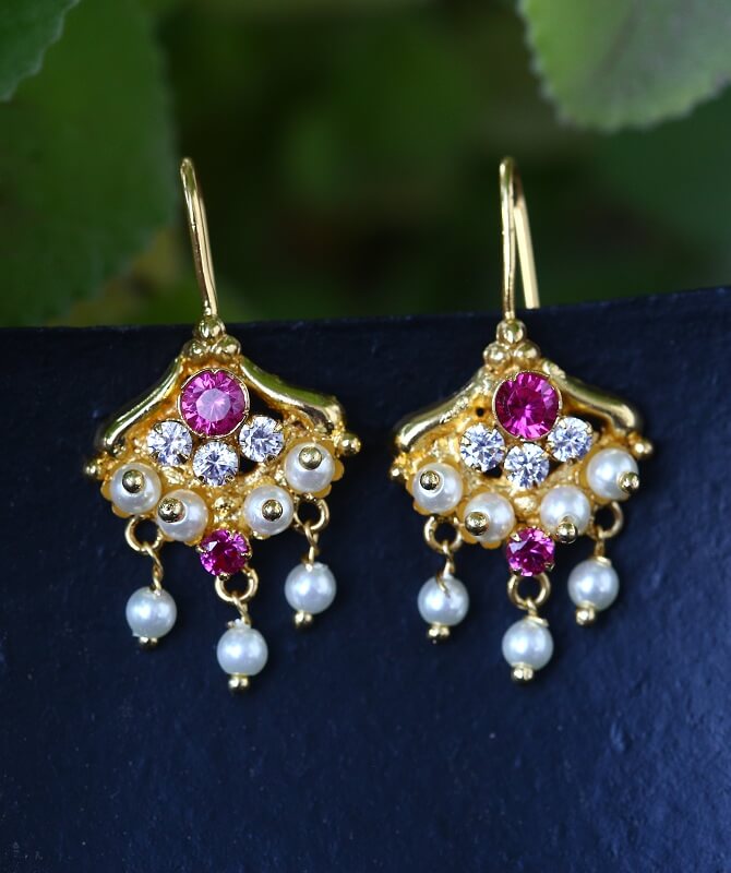 Beautiful golden earrings with sparkling pink gems, pearls, and diamonds displayed on a navy background against a blurred natural greenery backdrop.