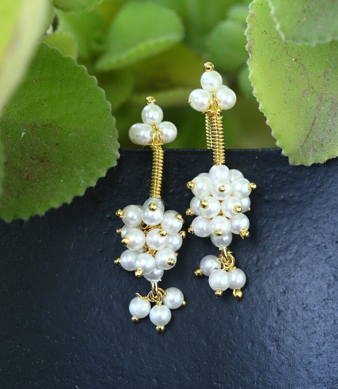 Elegant pearl cluster earrings from Mugdha Jewellery Store, featuring delicate white pearls arranged in a floral design on a gold-tone metallic frame, set against a backdrop of lush green foliage.