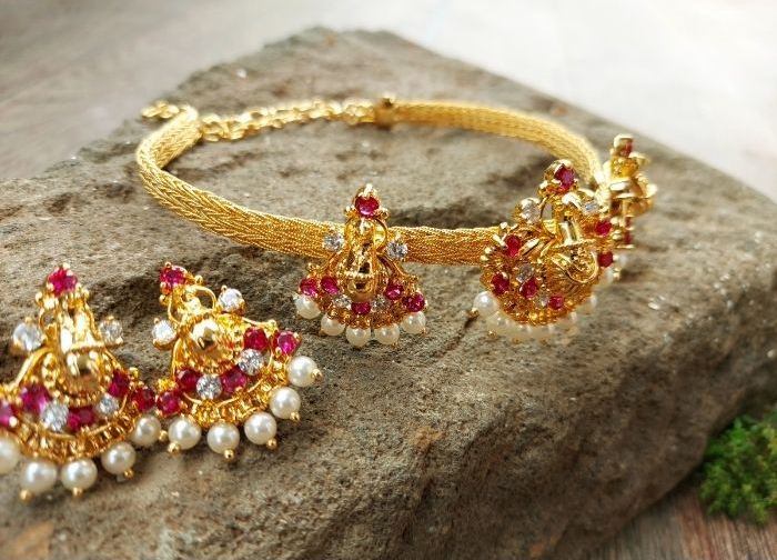 Opulent Peacock Bajuband and Choker Set: Enchanting gold-toned jewelry adorned with vibrant red gemstones and lustrous pearls, displayed on a rugged natural backdrop.