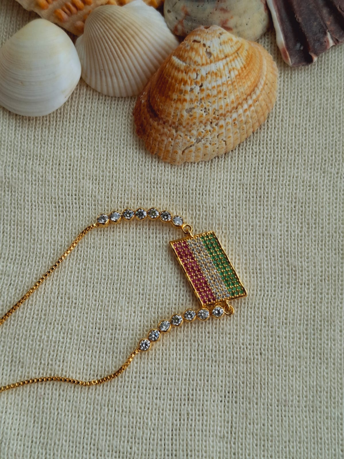 Elegant golden designer bracelet with colorful gemstones, displayed amidst seashells on a neutral fabric background from Mugdha Jewellery Store.