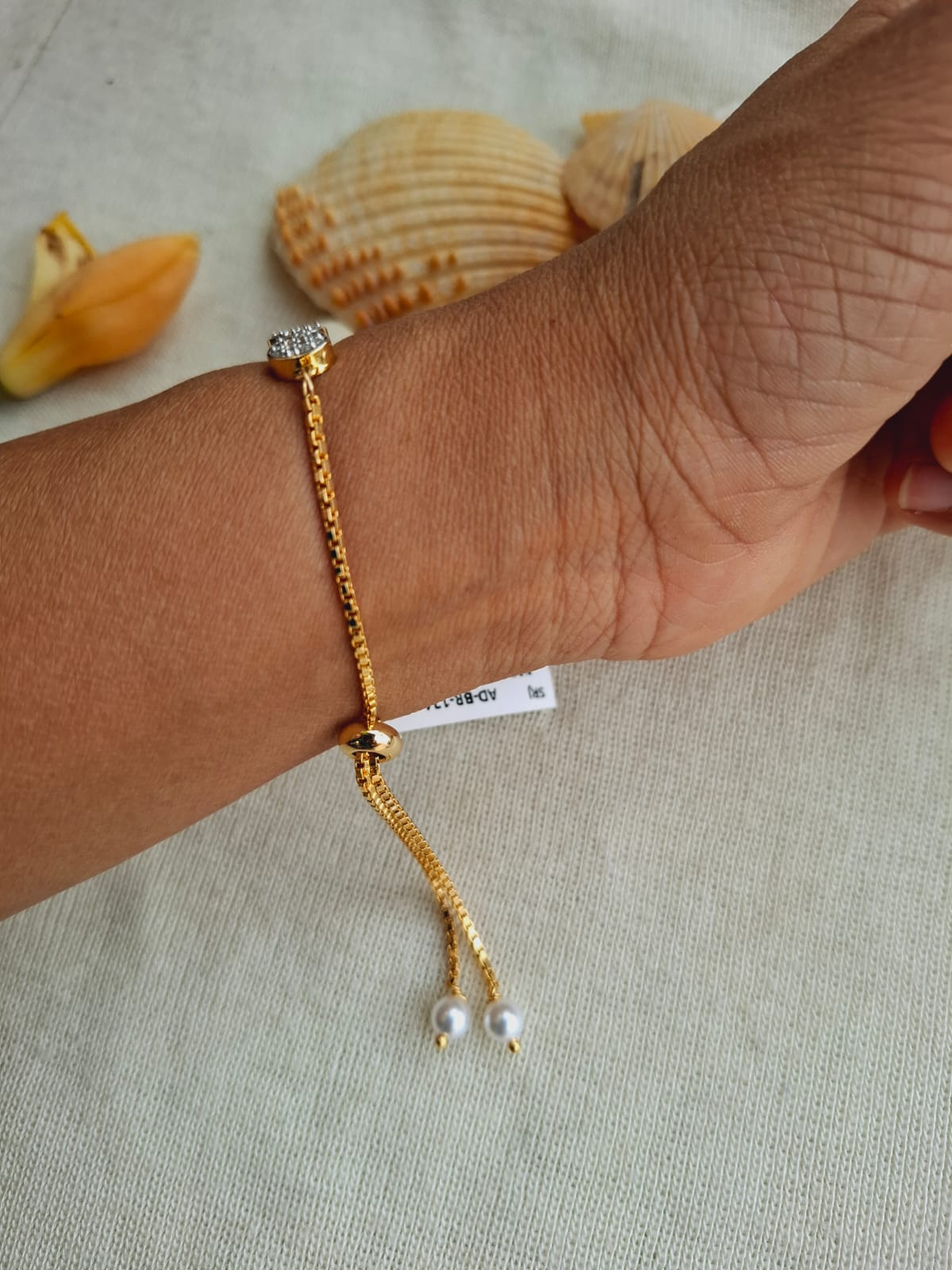Elegant gold-toned adjustable bracelet with dangling pearls, displayed on a human hand with shells in the background, from Mugdha Jewellery Store.