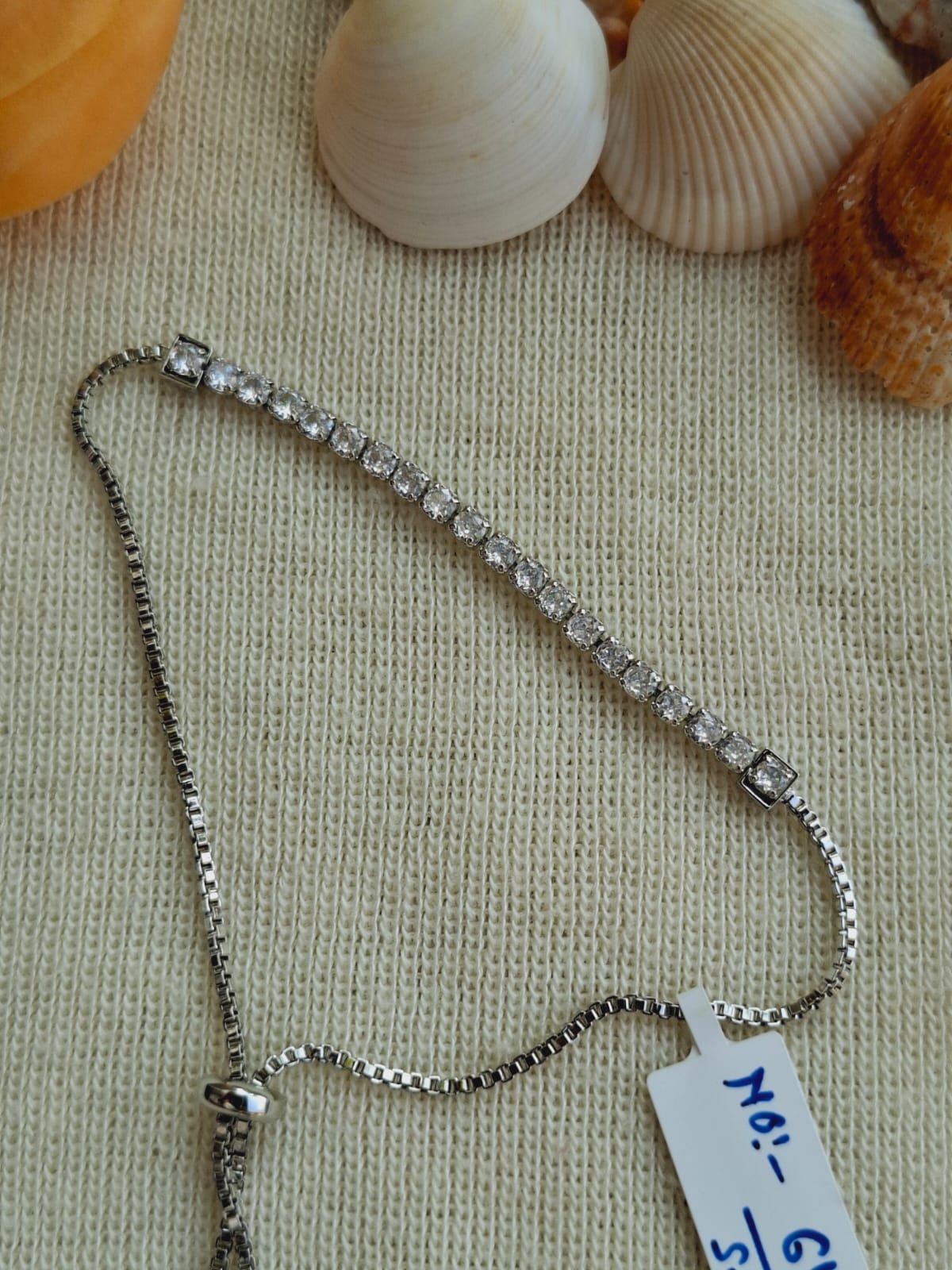Elegant Silver-Toned Adjustable Bracelet with Sparkling Rhinestones, Displayed on a Textured Fabric Background Alongside Seashells.
