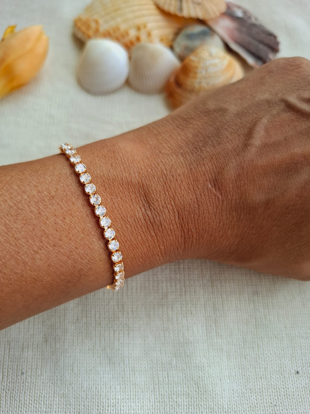 Elegant golden bracelet with sparkling American diamonds adorning a woman's wrist, set against a backdrop of seashells and beach-inspired elements.