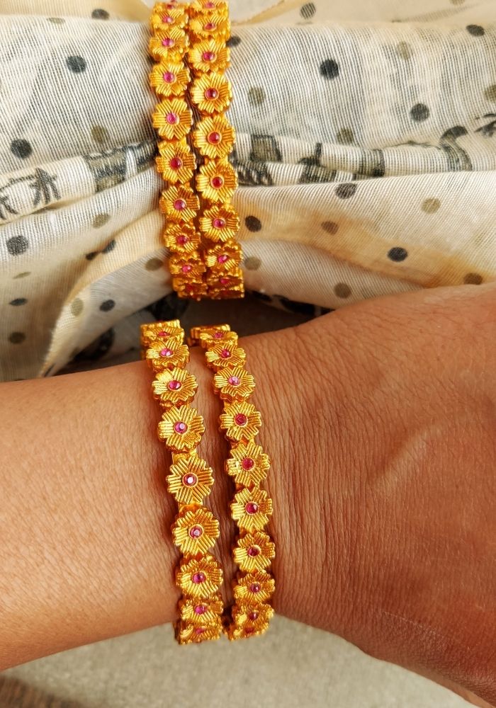 Ornate floral bangle bracelets on a woman's arm. The set of 4 gold-toned bangles are adorned with vibrant yellow and orange sunflower motifs, creating a stunning and eye-catching accessory. The bracelets are positioned against a patterned fabric background, adding to the overall stylish presentation.