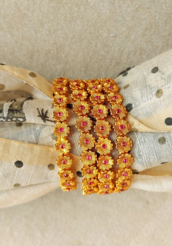 Four temple-style gold and red floral bangles stacked on a patterned fabric background in the Mugdha Jewellery Store product image.