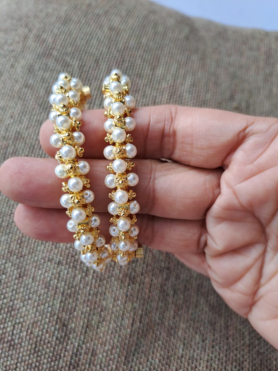 Traditional Gopi Tode. Set of 2
Two elegant gold-tone earrings adorned with delicate pearls, showcased on a hand against a textured gray background.