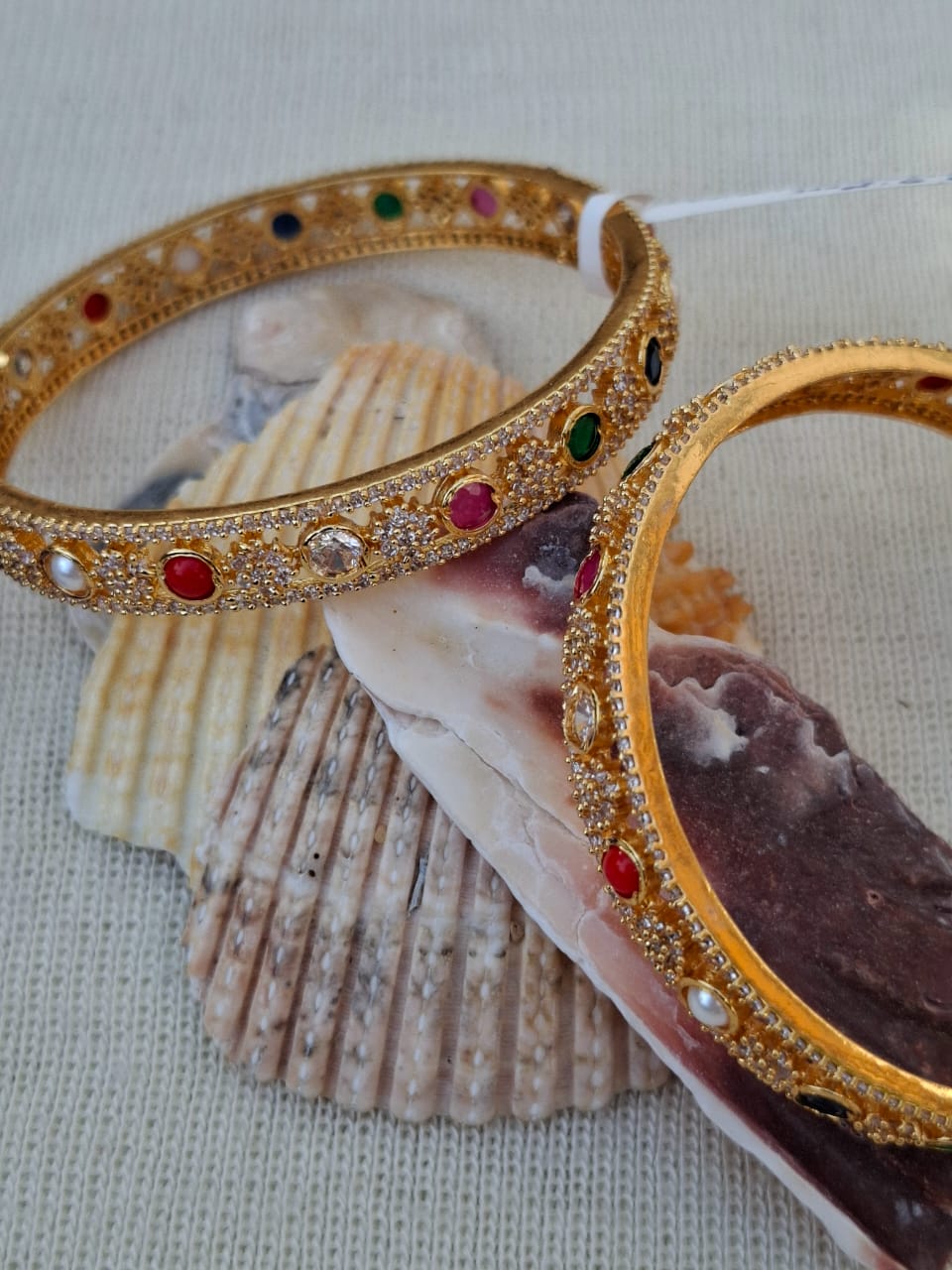 Ornate designer bangles set on seashell, showcasing intricate gold-tone metal work with colorful gemstone accents, displayed on a neutral background.