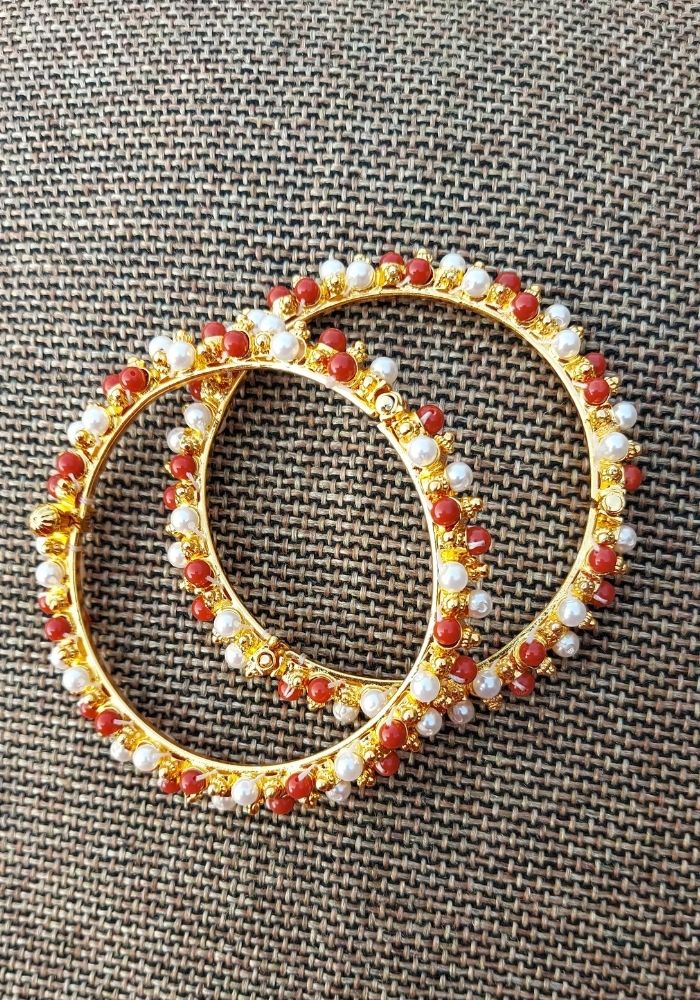 Pair of ornate gold metal bangles adorned with red and white gemstones, displayed on a textured gray fabric background.