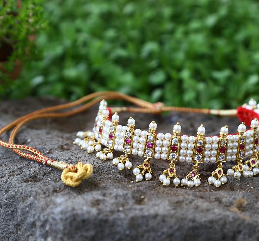 Ornate pearl and ruby studded gold choker necklace from Mugdha Jewellery Store, resting on a rocky surface against a lush green foliage background.