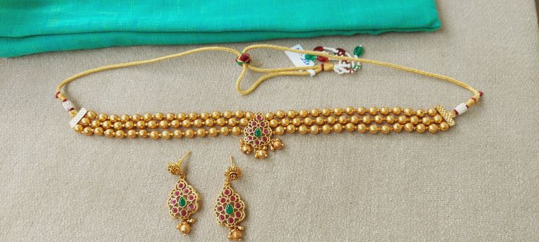 Gold beaded choker necklace with red and green gemstone pendant, paired with matching earrings from Mugdha Jewellery Store.