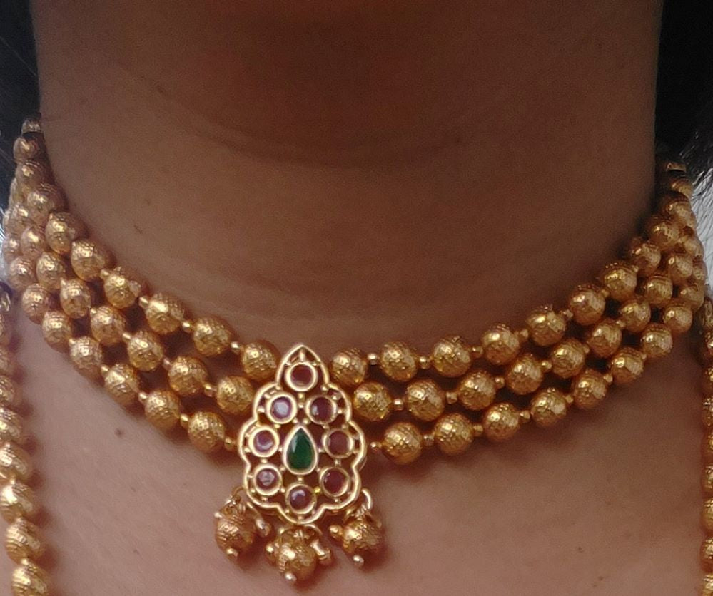 Golden beaded necklace with a center pendant featuring green and red gemstones, displayed on a plain background.