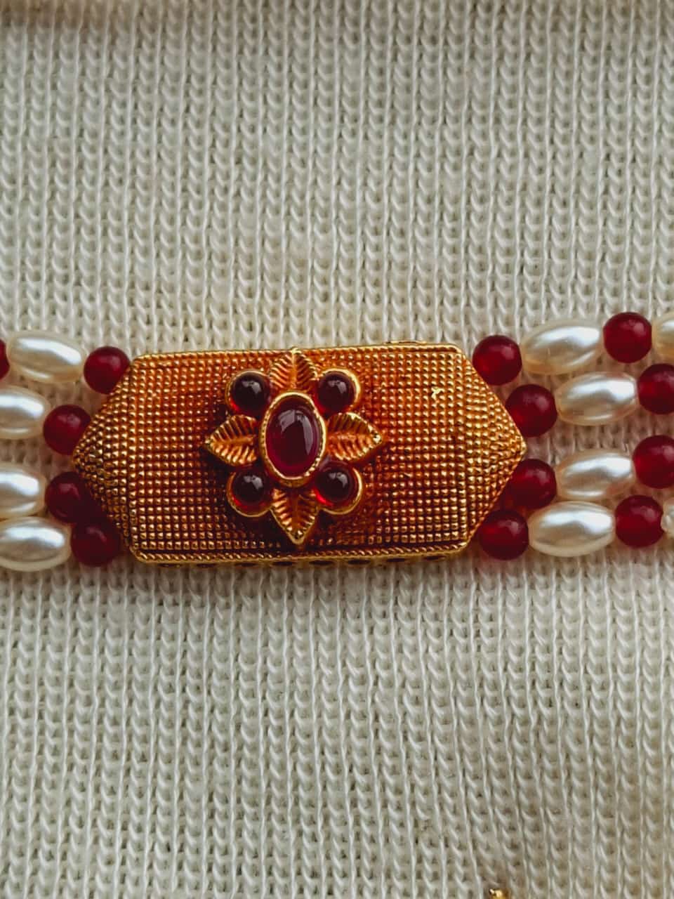 Rice Pearls Geru Designer Choker
A detailed golden choker with a floral centerpiece and red accents, surrounded by lustrous rice pearls against a beige knitted background.