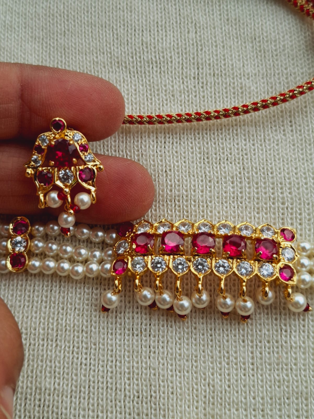 Elegant gold-toned choker necklace featuring a vibrant red and pearl pendant adorned with sparkling gems, showcased on a pearl-studded chain against a neutral fabric background.