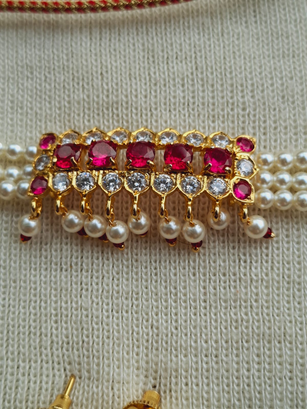 Detailed ornate choker necklace with rows of red gemstones and pearls, displayed on a light colored knitted fabric background.