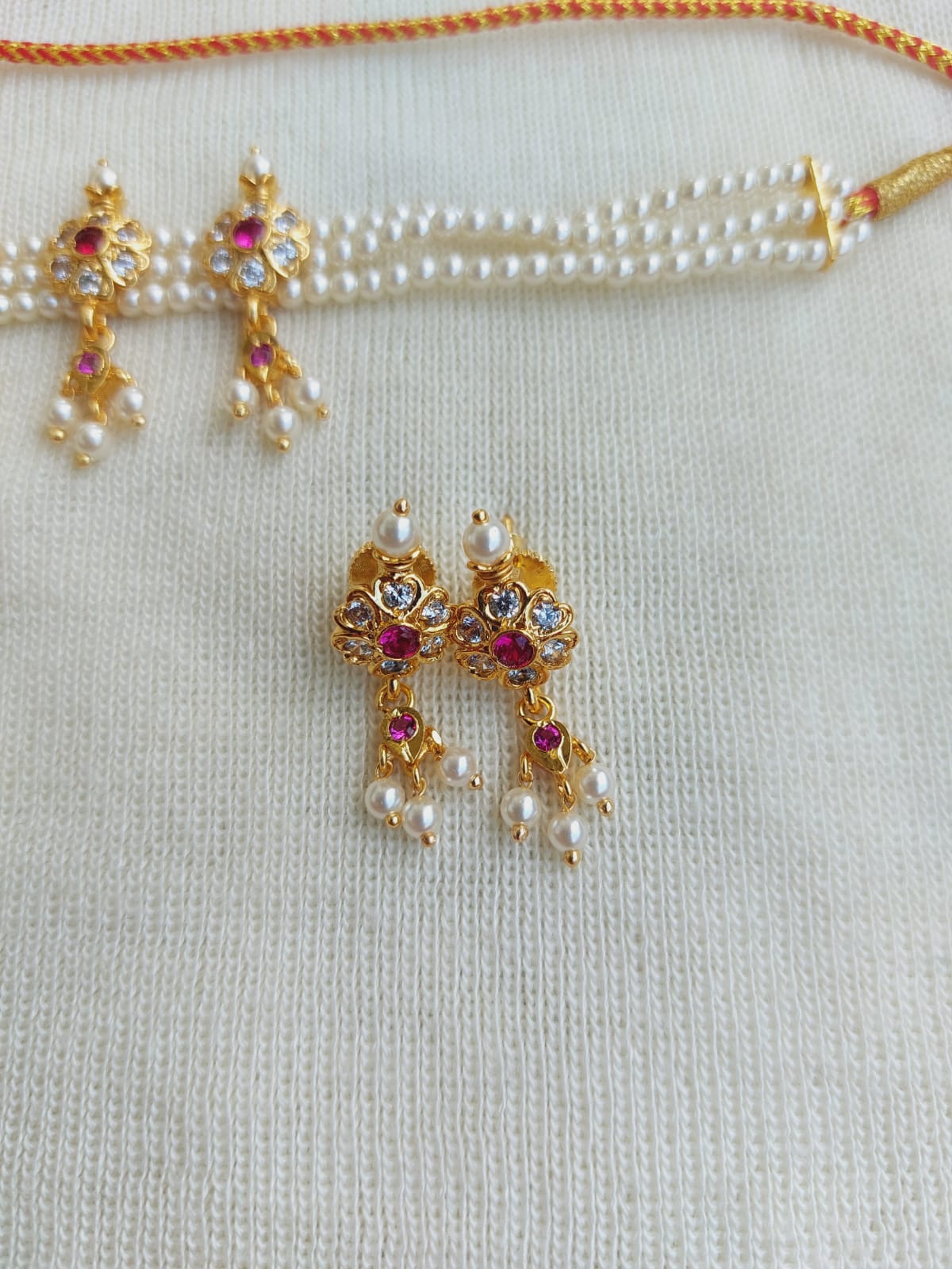 Elegant pearl and gold-toned floral-patterned necklace and earrings set from the Mugdha Jewellery Store. The necklace features a string of pearls accented with ruby-colored gemstones, while the earrings display intricate golden flower designs with cascading pearl details.