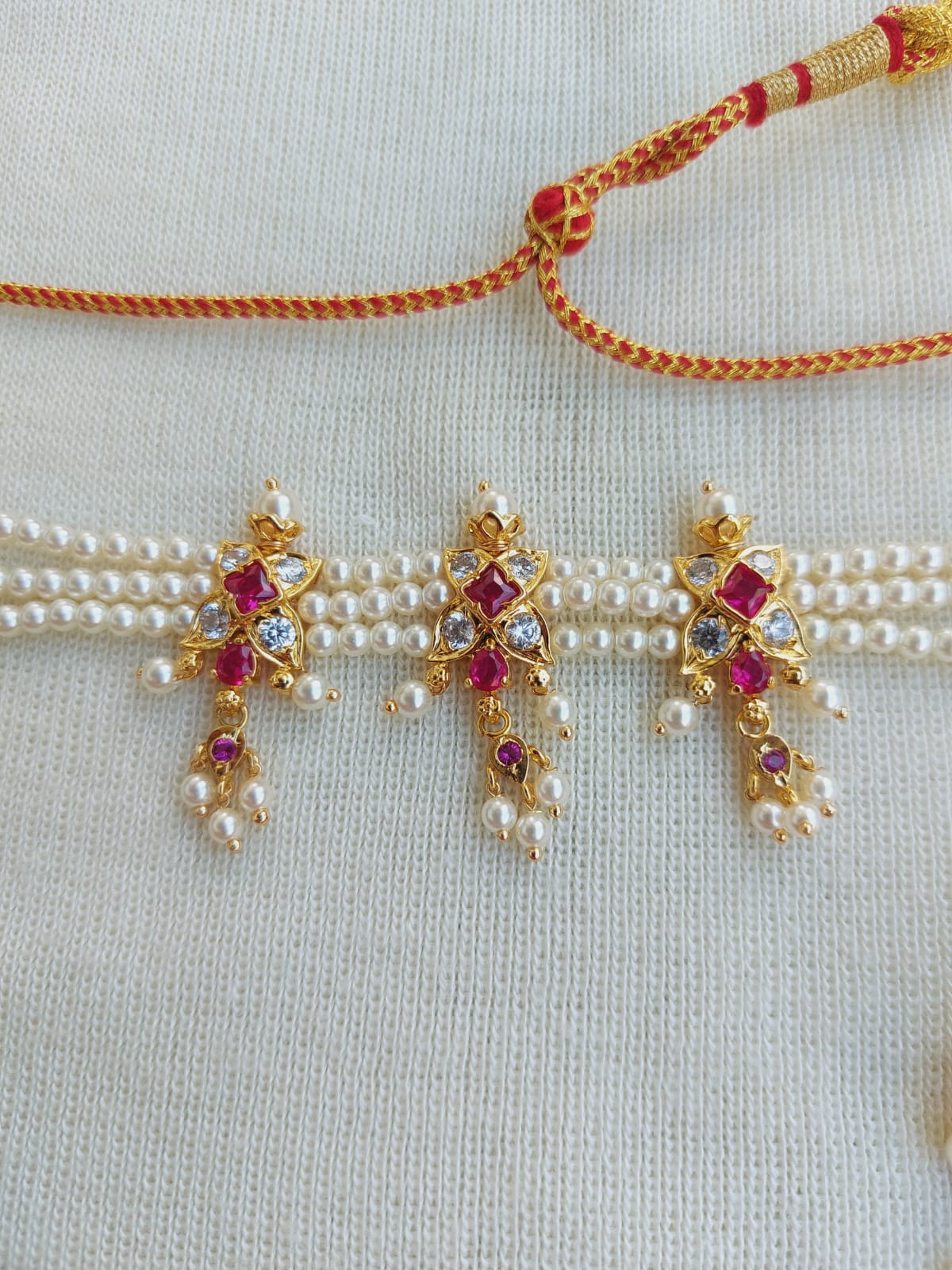 Beautiful 3 Patti Designer Red Flower Chinchpeti from Mugdha Jewellery Store. The image shows a delicate gold and pearl necklace with a distinctive red floral charm pendant, surrounded by a tasseled chain in vibrant orange.