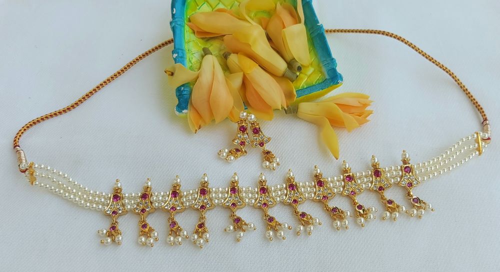 Elegant gold-tone necklace with intricate floral design, featuring a string of pearls and sparkling rhinestones, showcased against a vibrant backdrop from Mugdha Jewellery Store.