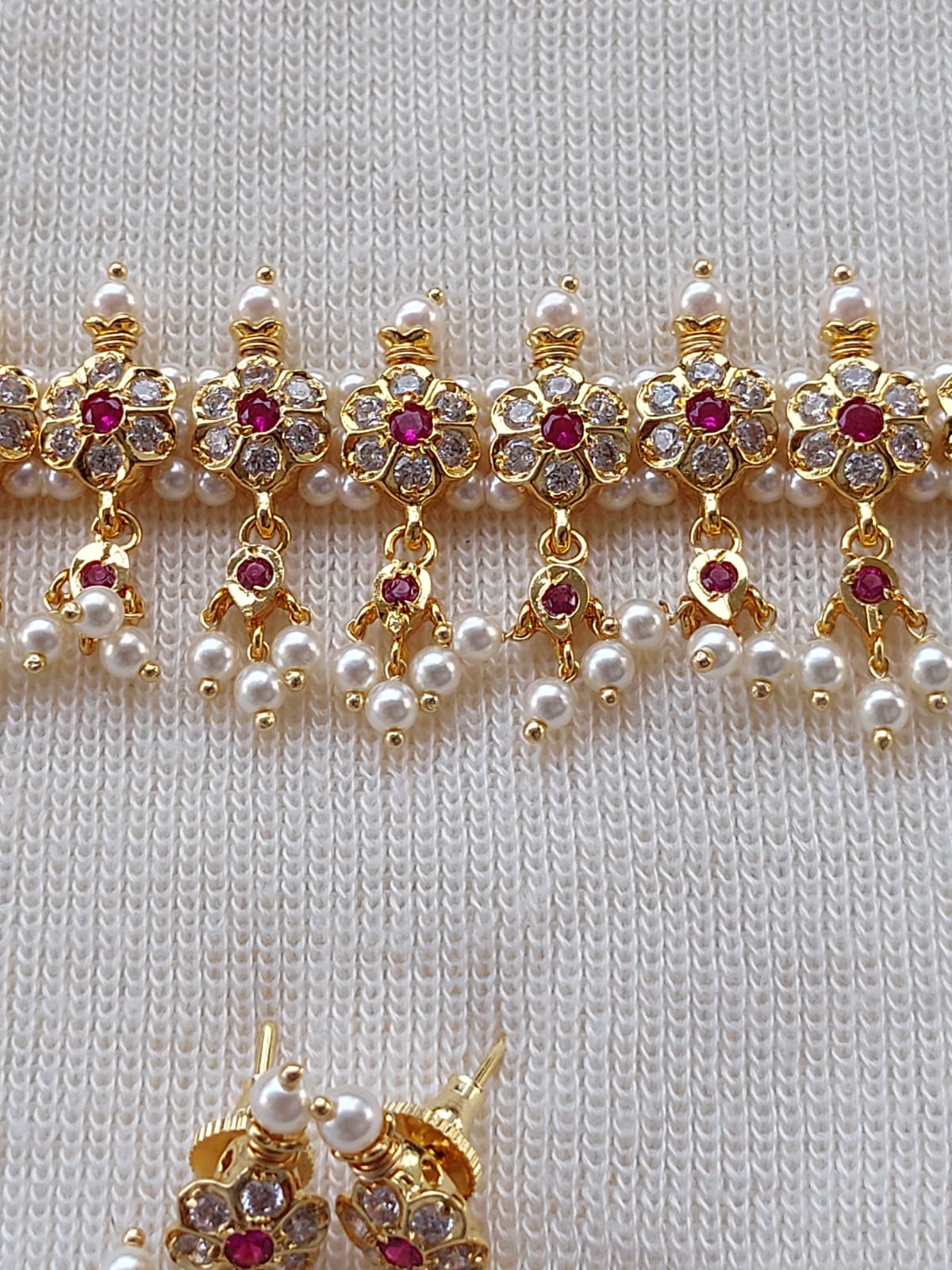 Ornate gold-toned flower-shaped pendant with red and white gemstone accents, set against a cozy cream-colored knit fabric background.