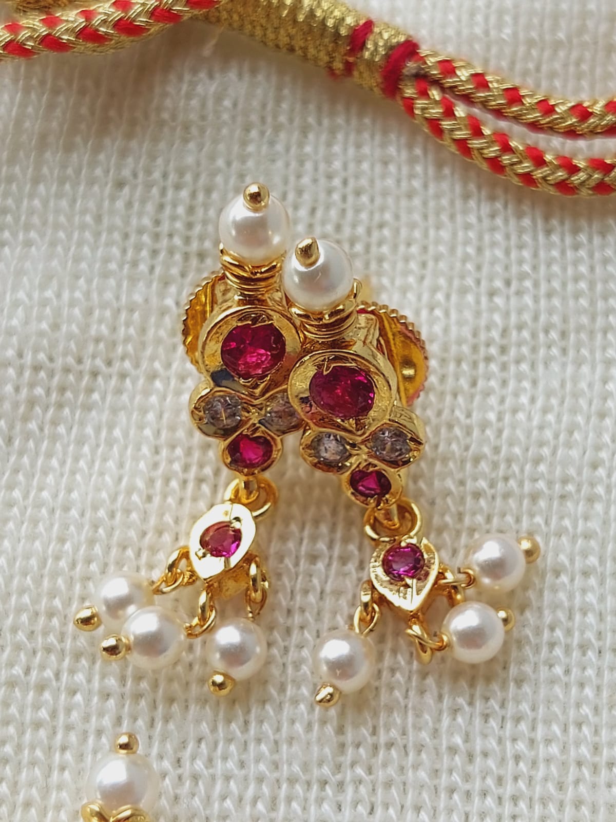 Gold-toned ornate earrings with red gemstones and white pearls, displayed against a white knitted fabric.