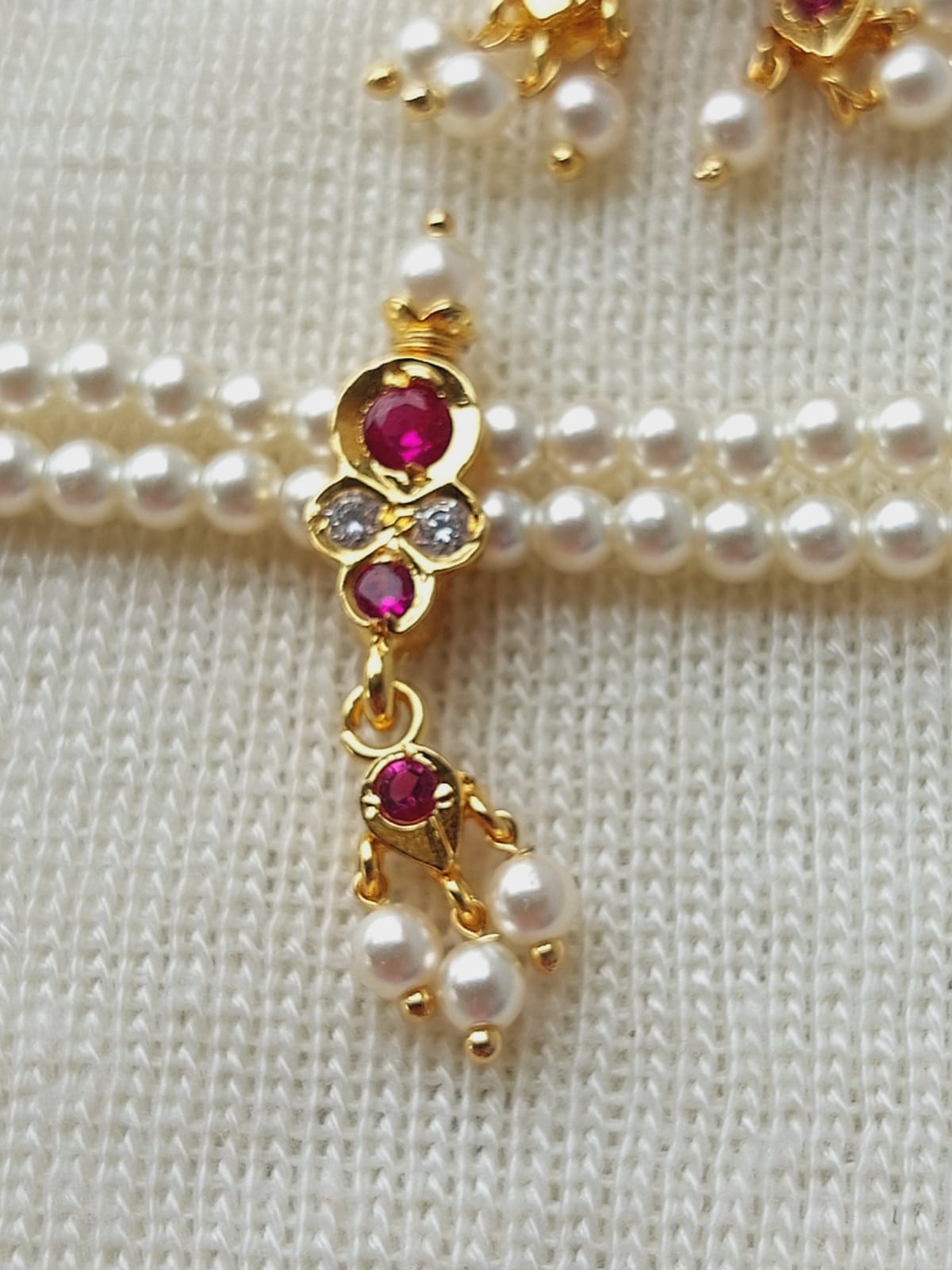 Elegant gold pendant with rubies and diamonds, hanging amidst a strand of lustrous pearls on a cream knitted fabric background.