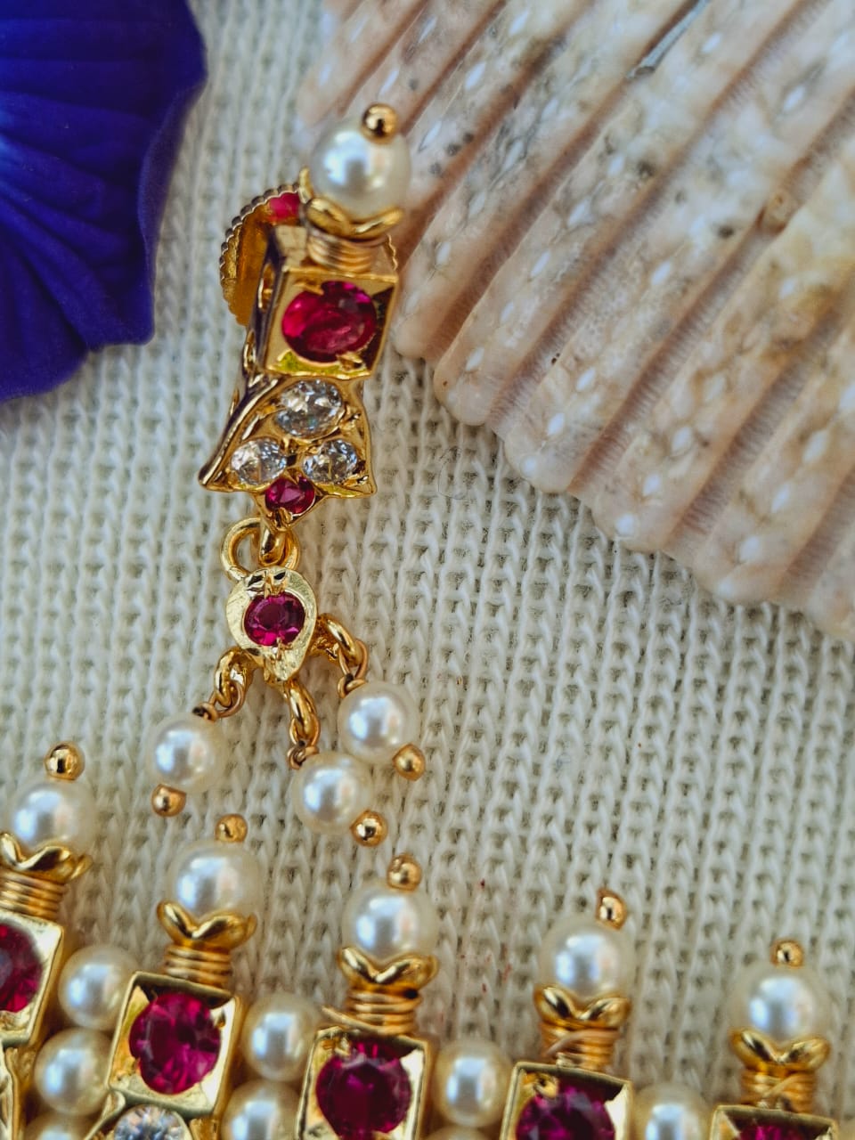 Ornate gold-toned chinchpeti (headpiece) adorned with vibrant red gemstones, white pearls, and intricate detailing, displayed on a textured fabric backdrop.