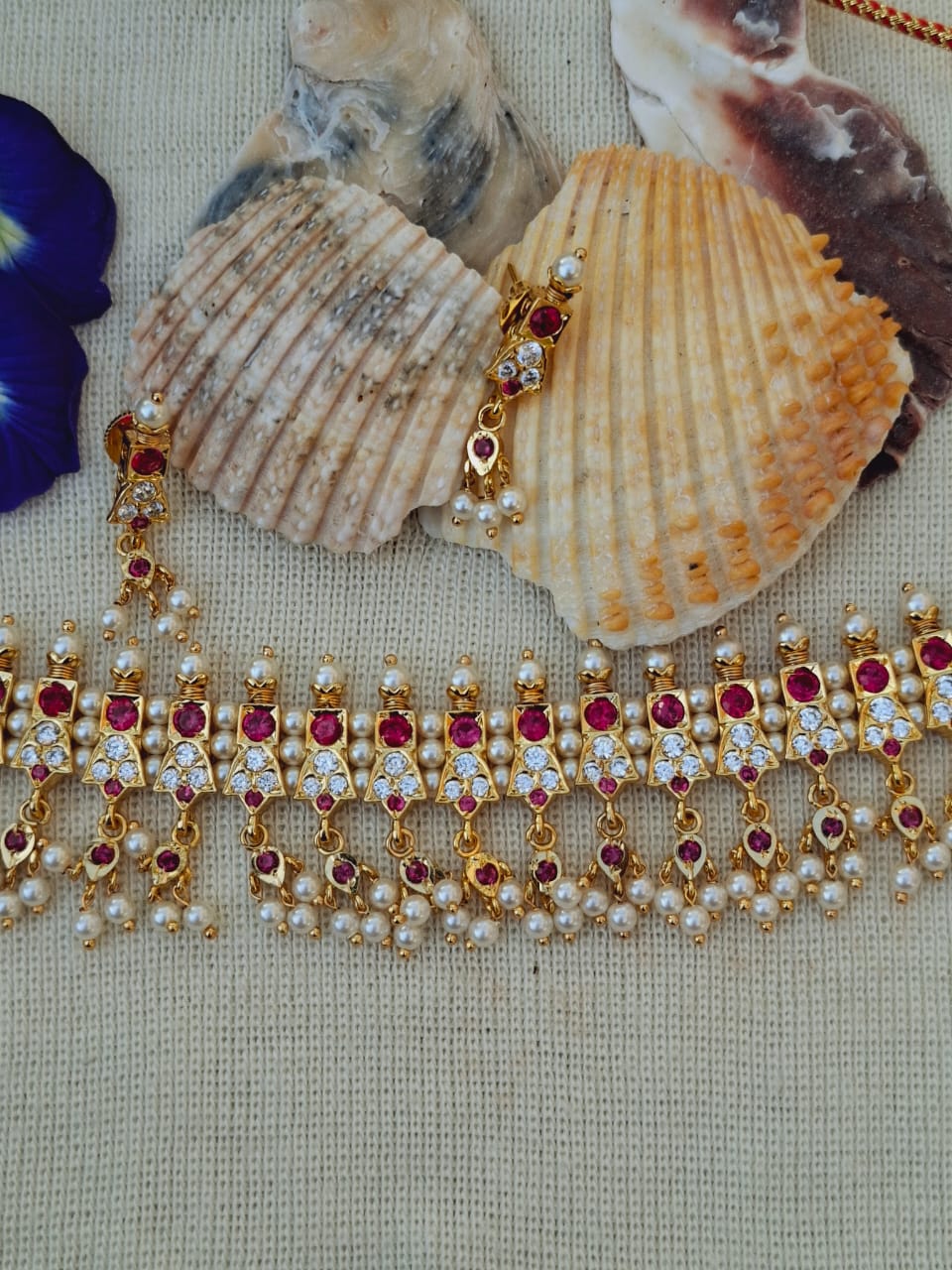 Ornate gold and ruby necklace with shell accents, showcased on a textured background in the Mugdha Jewellery Store.