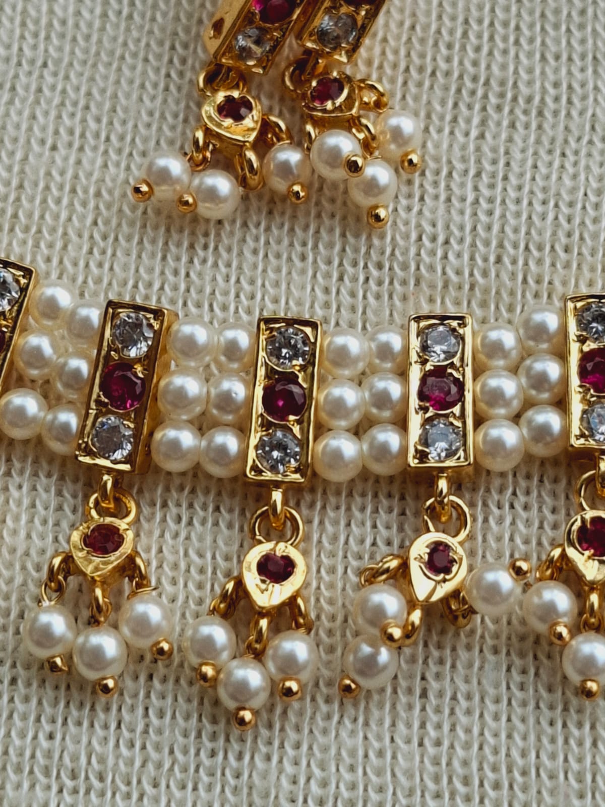 Gold-tone traditional chinchpeti with white pearls, red gemstones, and rhinestones on a knitted background.