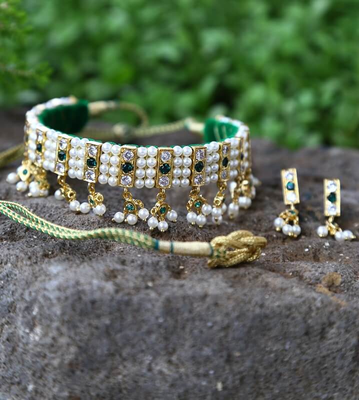 Ornate green and gold jeweled choker necklace with dangling pearls, displayed on a rocky, natural background.