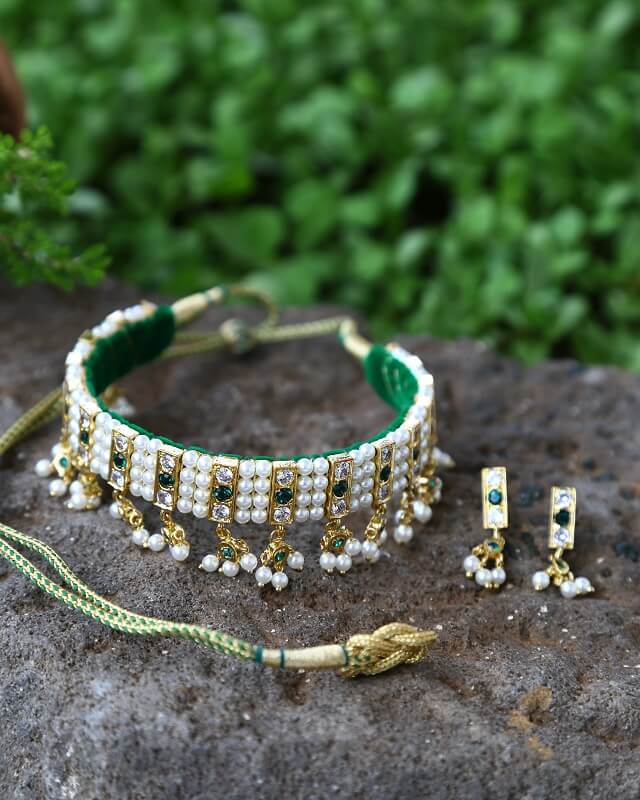 Green White Stone 4 Patti Gadi Chinchpeti
A beautiful green and white stone studded choker necklace with a tassel pendant, paired with matching stud earrings, displayed on a natural stone surface against a blurred green foliage background.