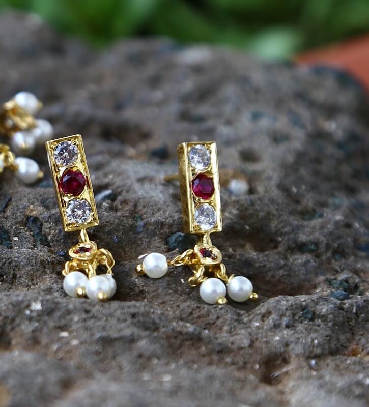 Sparkling gold-tone earrings with red and white stones, featuring dangling pearl accents, resting on a rocky surface with a natural blurred background.