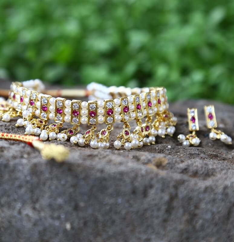 23 Patti Dark Pink White Choker Chinchpeti with pearl and ruby-encrusted gold toned jewelry set placed on a rocky surface against a blurred green natural background