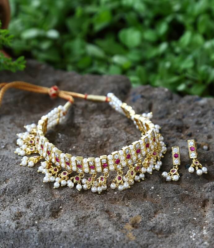 Elegant dark pink and white jewellery set with intricate design and pearl accents, displayed on a rocky surface with a lush green natural background.