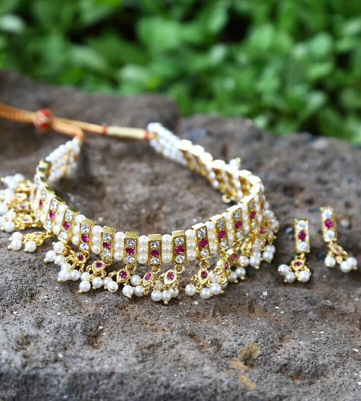 23 Patti Dark Pink White Choker Chinchpeti Necklace and Earrings Set
A stunning gold-toned choker necklace with intricate dark pink and white gemstone detailing, displayed on a rocky surface with lush greenery in the background.