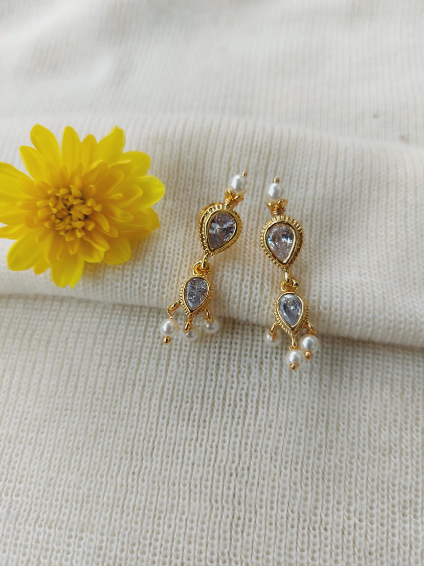 Elegant gold-tone earrings with sparkling stones and dangling pearls, showcased on a white knit background with a vibrant yellow flower.