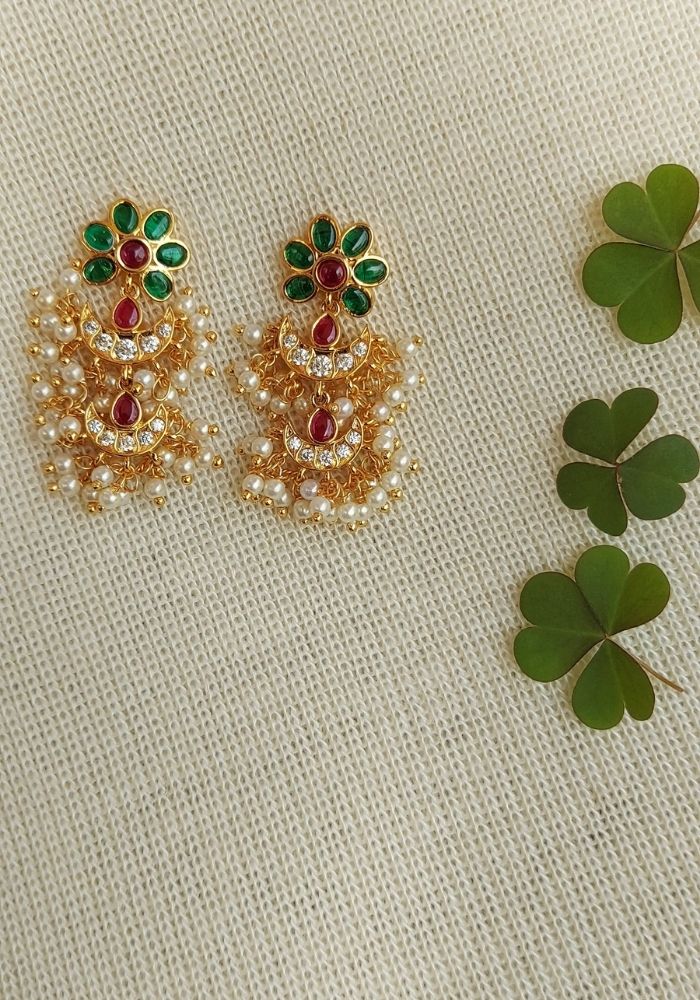 Glamorous gold, pearl, and gemstone earrings from Mugdha Jewellery Store. Intricate floral design with green, red, and yellow stones set in an ornate cluster. Delicate and sophisticated accessory displayed on a textured white background with lush green shamrocks.