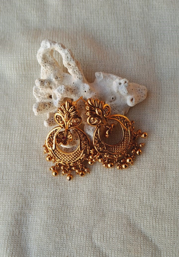 Elegant gold-toned dangler earrings with intricate carved designs, featuring eye-catching patterns and hanging beaded accents, displayed on a natural rock formation.