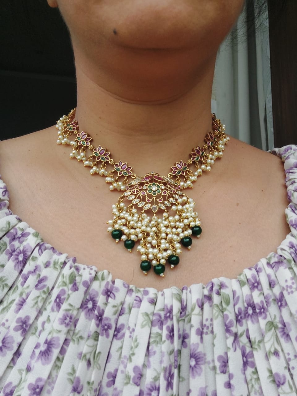 Ornate golden lotus necklace with intricate floral patterns, pearls, and green gemstones, displayed prominently against a vibrant floral dress.