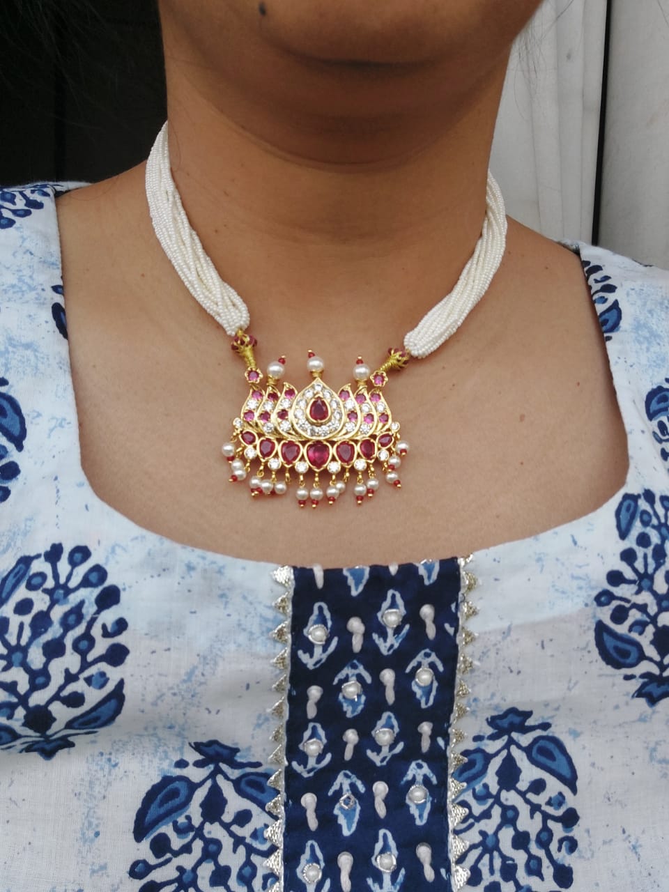 Elegant pearl necklace with intricate gold and ruby pendant, worn with a patterned blue and white blouse.