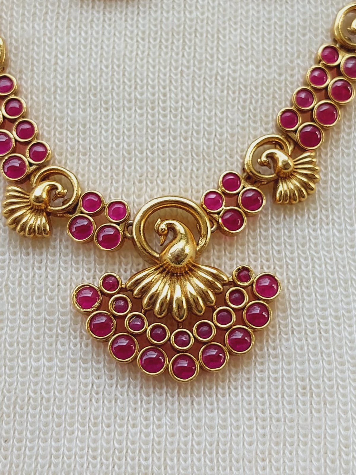 Ornate gold necklace with vibrant ruby gemstones, showcasing a centered peacock design surrounded by swirling floral motifs, set against a light colored knitted fabric background.