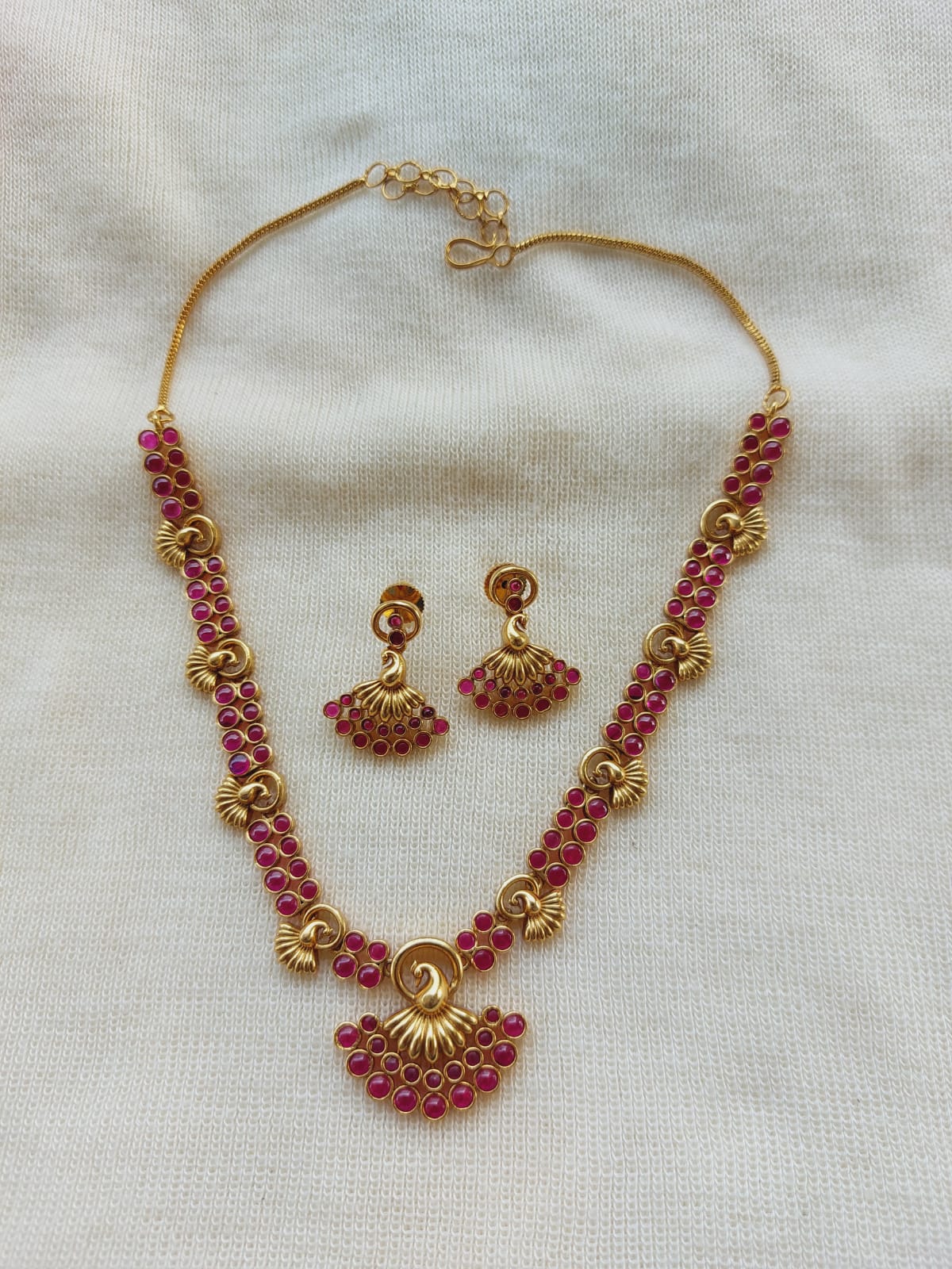 Elegant gold and ruby necklace with matching earrings featuring intricate peacock-inspired design, displayed on a neutral background.