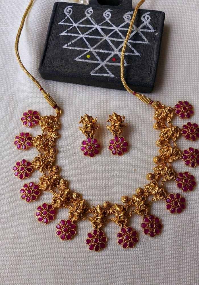 Ornate gold floral necklace with vibrant red gemstones, displayed alongside a black textile with intricate geometric patterns, showcased against a plain white background.