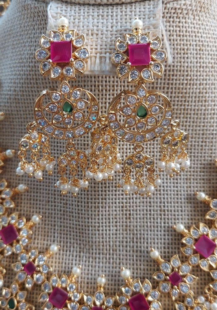 Elegant gold-toned necklace and earrings set from Mugdha Jewellery Store, adorned with vibrant red gemstones and sparkling crystal embellishments, creating a regal and eye-catching look.