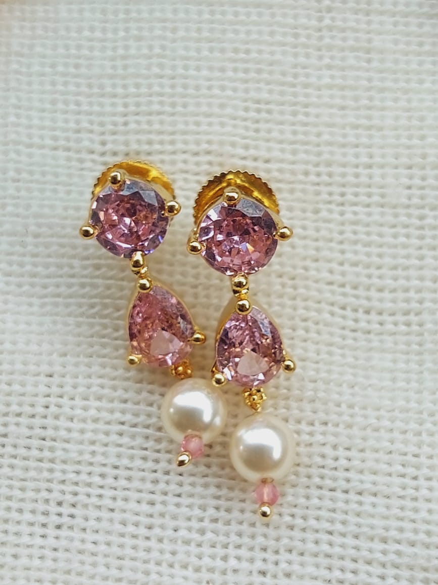 Elegant gold-toned earrings with light pink sparkling stones and delicate white pearls, displayed on a textured fabric background.