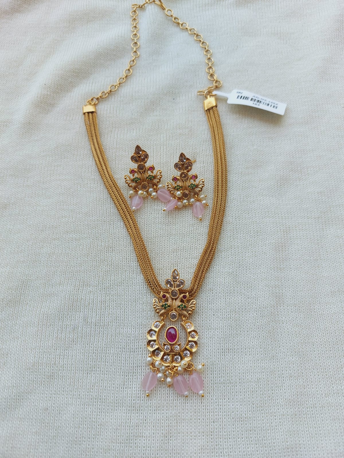 Elegant designer peacock-inspired kundan jewelry set with baby pink beads, featuring a pendant necklace and matching earrings, showcased on a white fabric background.
