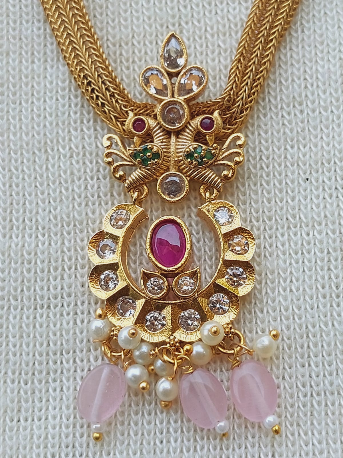 Ornate designer peacock-inspired kundan necklace set with pink beads, precious stones, and pearls on a white knitted background.
