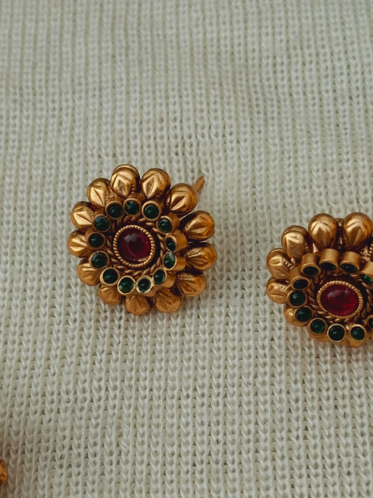 Ornate golden floral earrings with ruby and emerald gemstones displayed on a white knitted fabric background.