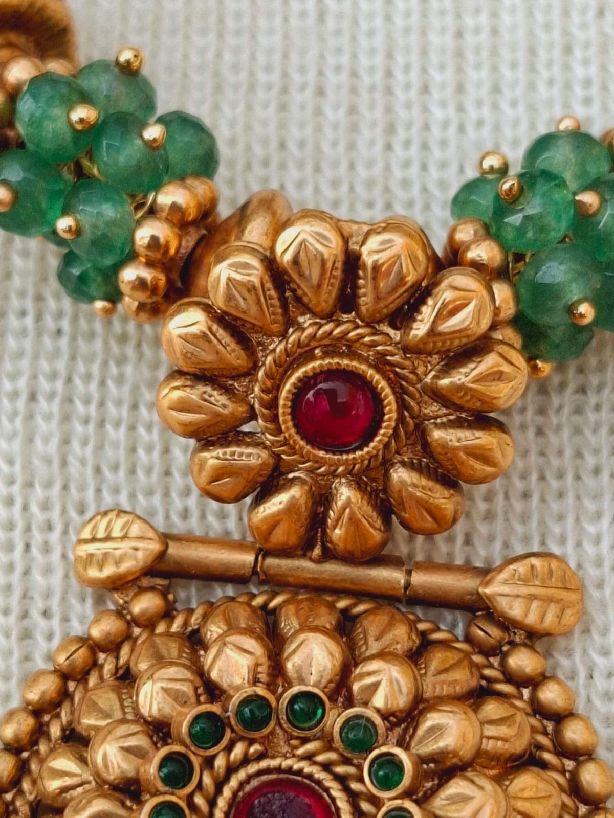 Ornate gold and green gemstone designer necklace from Mugdha Jewellery Store featuring intricate floral motifs and a central ruby-like gemstone.