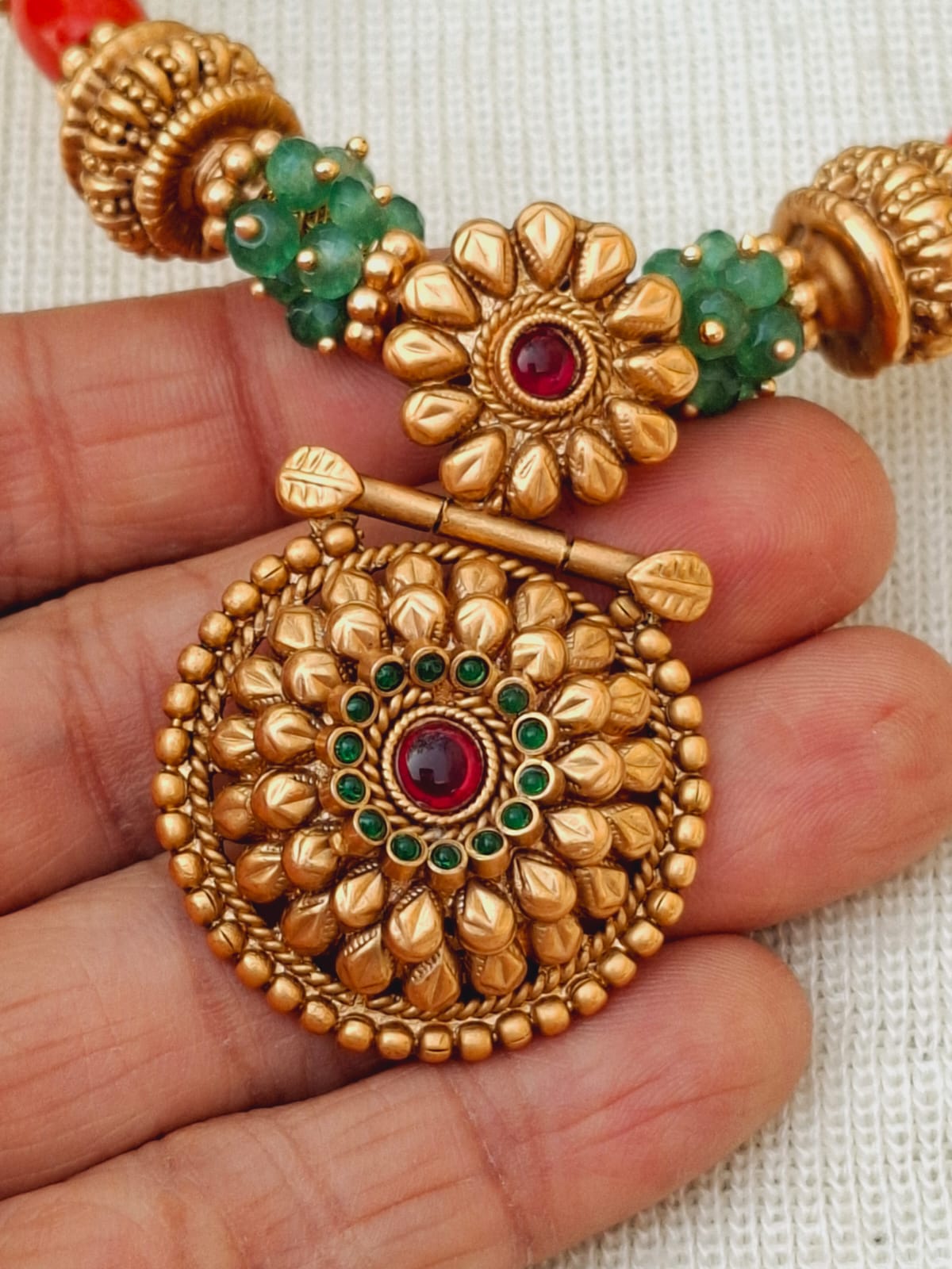 Ornate gold-toned necklace with intricate flower-shaped pendants adorned with red and green gemstones, displayed on a white textured surface.